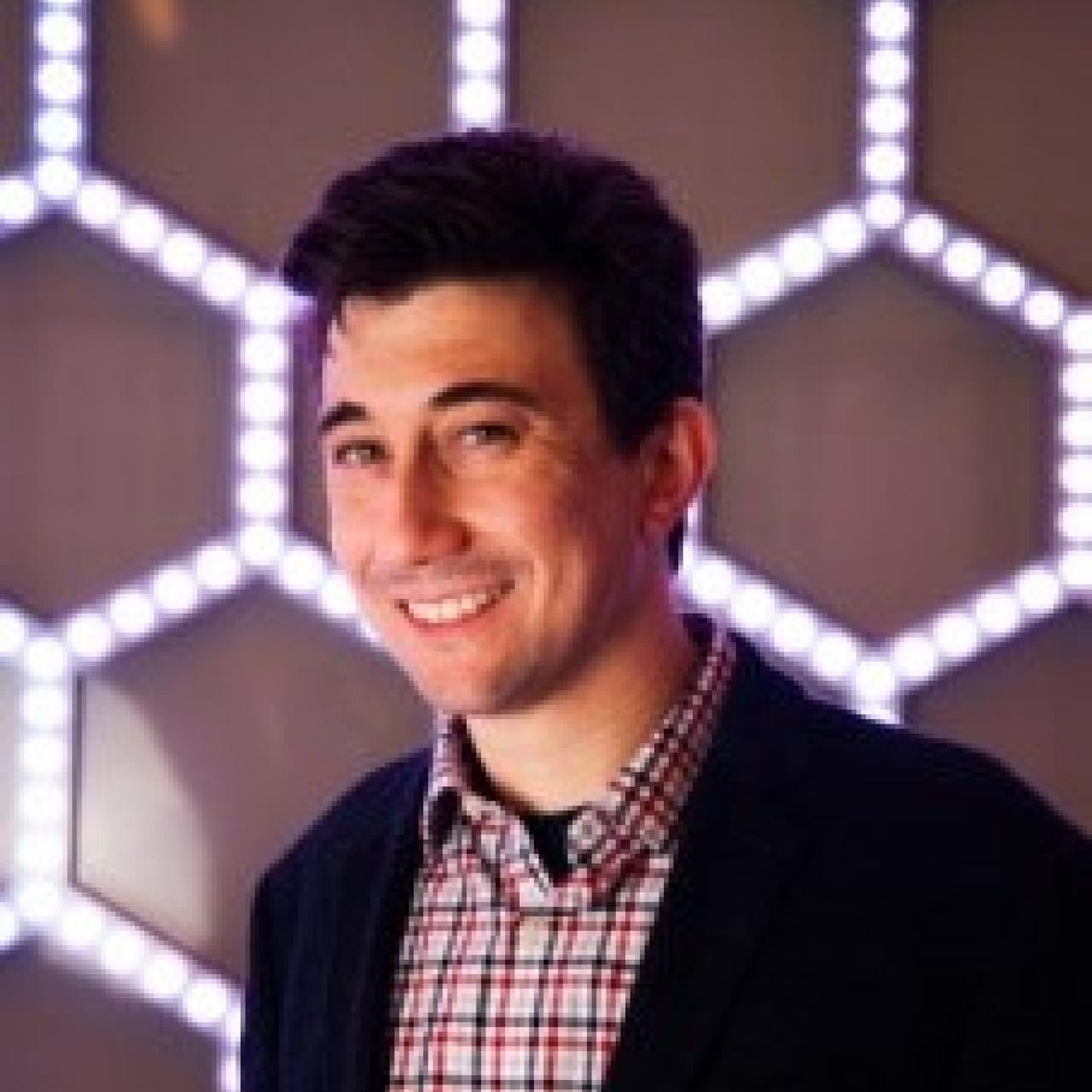 A joshua goldberger poses in front of a hexagonal patterned backdrop that reflects scientific themes in materials science.