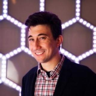 A joshua goldberger poses in front of a hexagonal patterned backdrop that reflects scientific themes in materials science.
