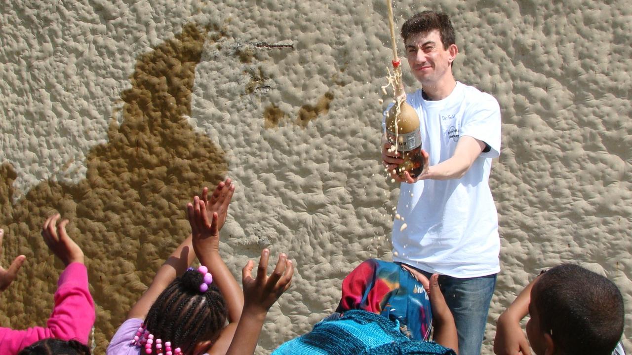 A demonstrator conducting a science experiment with a group of excited elementary school students reaching up to observe.