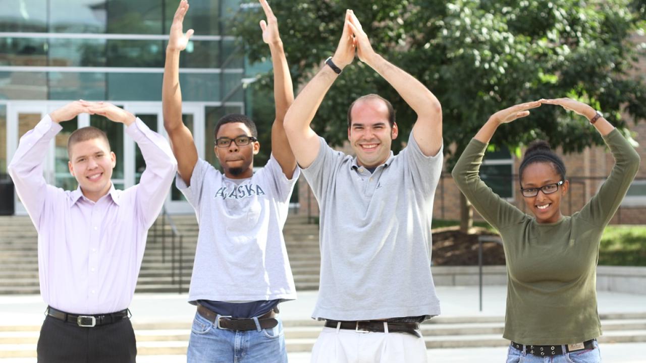 The image shows four students standing in front of a building, all raising their arms above their heads in a similar celebratory or playful pose. They are smiling and appear to be on a college campus, with steps and glass building facades visible in the background.